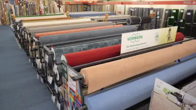 Mehrere aufgerollte Teppichbodenbeläge stehen in geordneten Reihen im Lagerbereich von TTL Heilbronn. Sichtbar sind verschiedene Farben, Muster und Materialien sowie ein Vorwerk-Hinweisschild zwischen den ausgestellten Bodenbelägen.