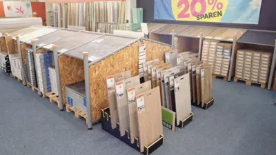 Innenansicht eines Fachgeschäfts mit zahlreichen Teppich- und Bodenbelagsmustern auf Paletten und in aufrechten Präsentationsständern. Im Hintergrund sind Stoff- und Materialproben sowie ein Banner mit einem Rabattangebot von 20 Prozent zu sehen.