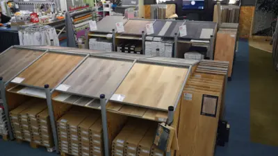 Auswahl an Laminat- und Holzdekorböden in einem Fachmarkt. Mehrere Musterplatten liegen auf Präsentationstischen, während verpackte Bodenbeläge darunter und in Regalen im Hintergrund ausgestellt sind.
