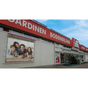Außenansicht eines Fachmarktes für Heimtextilien mit roter Beschilderung, Eingangsbereich und großformatigem Werbeplakat an der Gebäudefassade. Der Markt steht unter blauem Himmel und ist von einer gepflasterten Fläche umgeben.